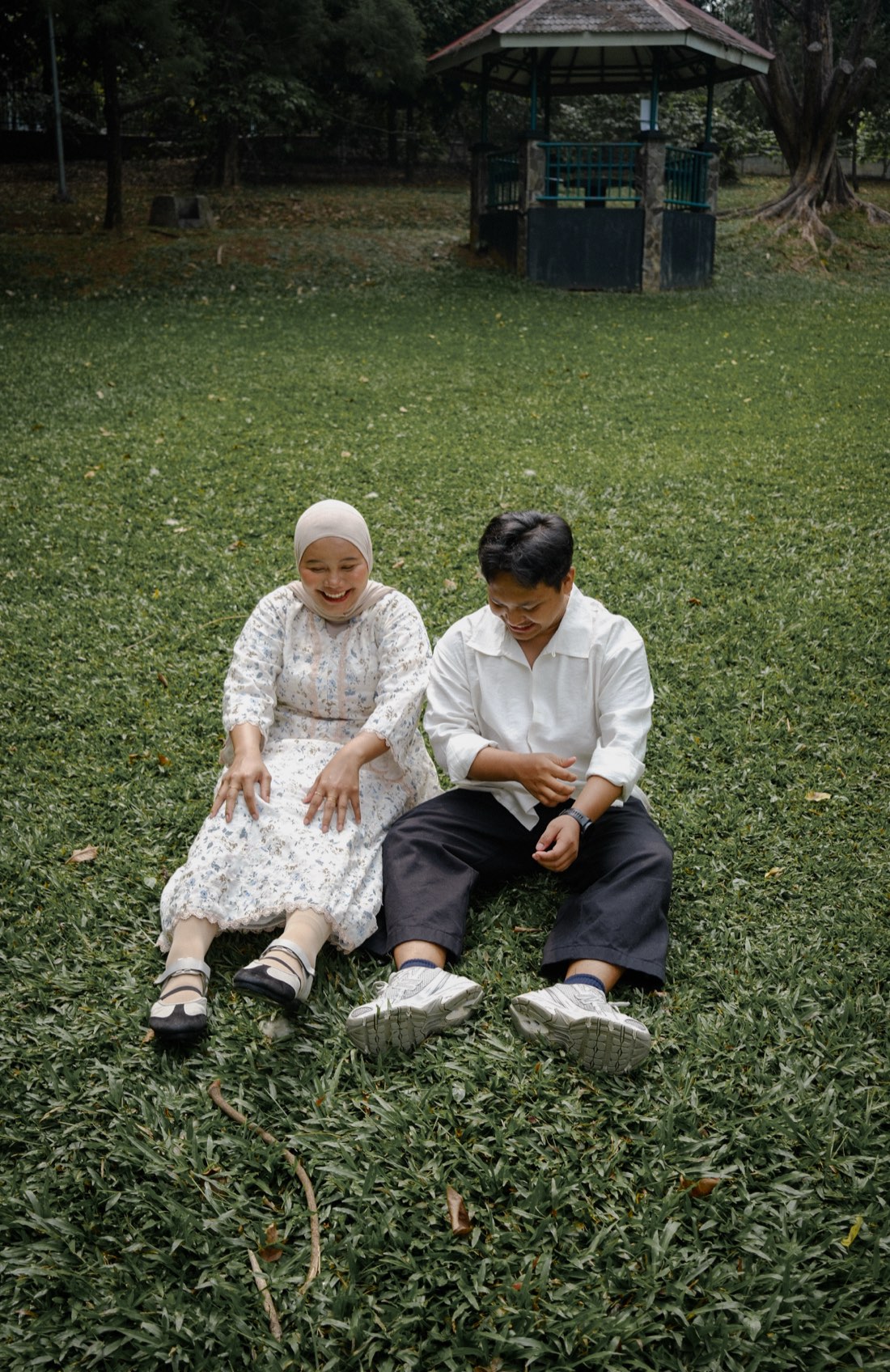 Deffy and Iqbal sitting side by side on the grass, laughing as they look down.