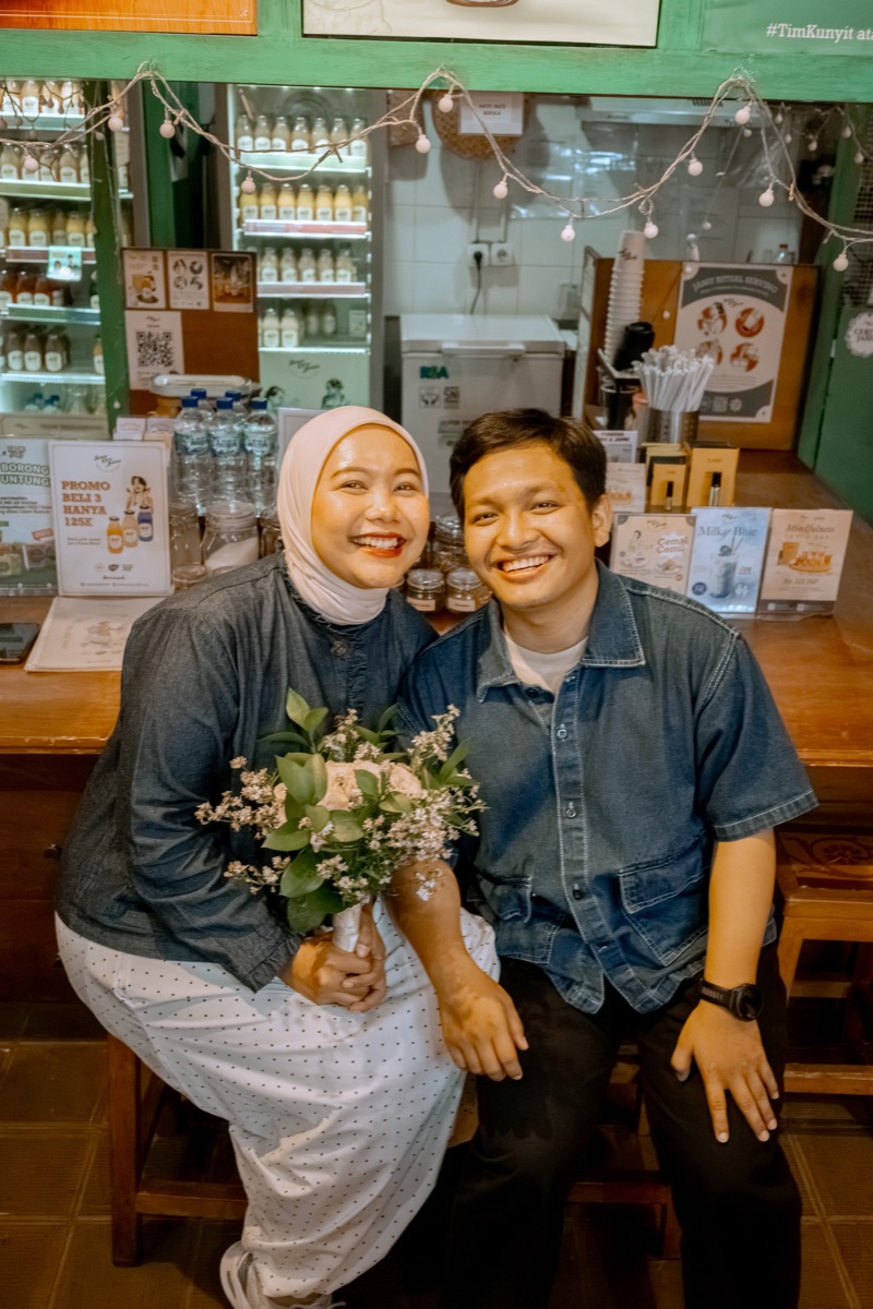 Iqbal and Deffy sitting close at the herbal drink counter, Deffy holding a small bouquet.