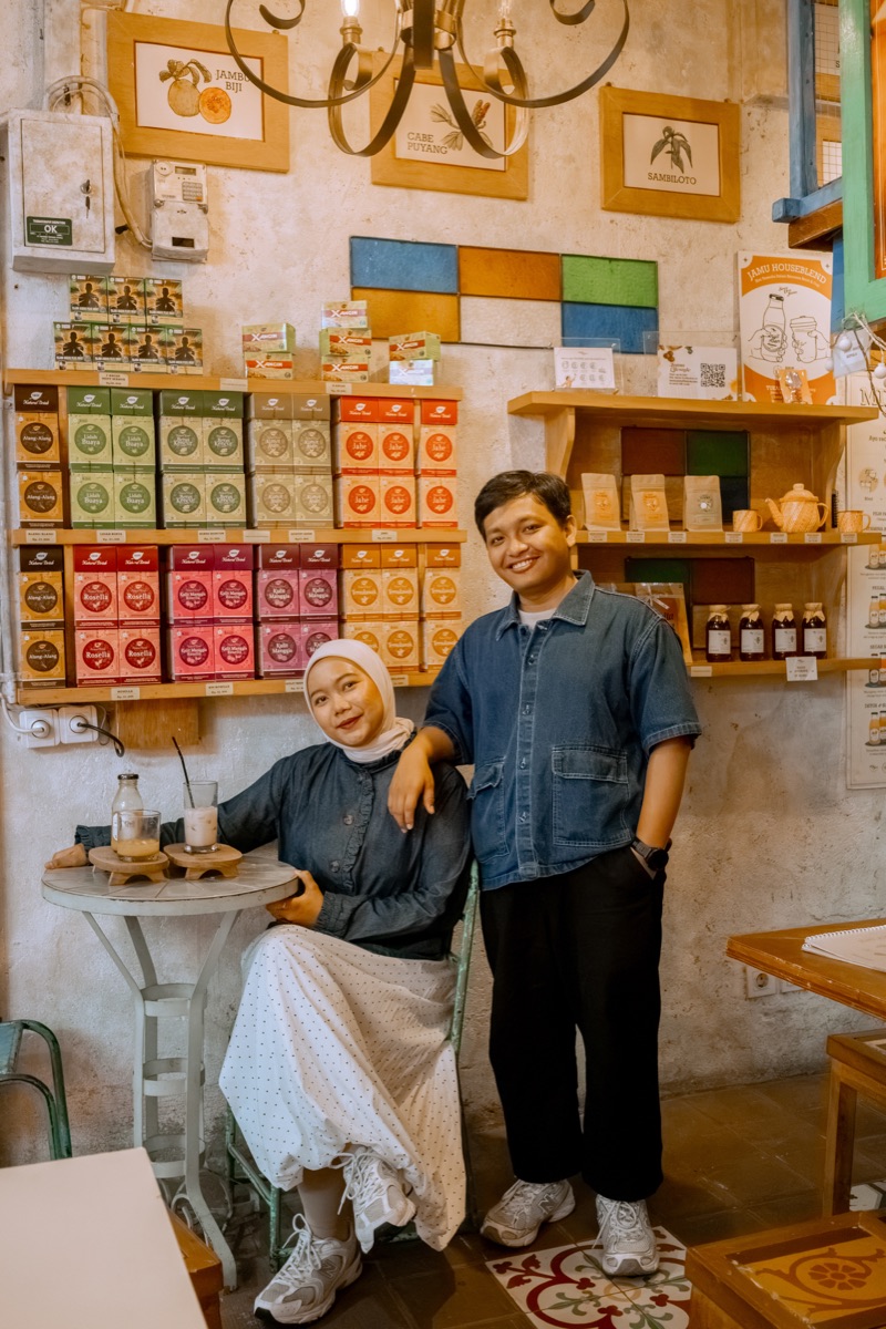 Deffy seated at a small table with Iqbal leaning beside her, in front of shelves of herbal drinks.